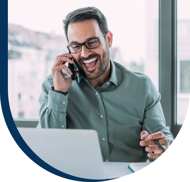A smiling man talking on a phone and looking at a laptop.